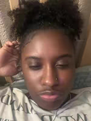 A close-up shot of a young Black woman with curly hair pulled back, wearing a gray t-shirt. Her eyes are mostly closed, and her lips are slightly pursed. A nose piercing is visible. The background is softly blurred, suggesting a casual indoor setting.