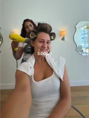 A selfie-style video shows a woman with hair rollers getting her hair styled. Another woman is behind her, holding a yellow hair dryer. The timestamp '5:00pm' is overlaid on the image.