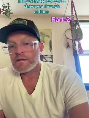 A man wearing glasses and a cap is in the foreground, facing the camera and speaking. The background is a room with decorations and a piece of art on the wall. Text overlays are present on the top and right sides of the image.