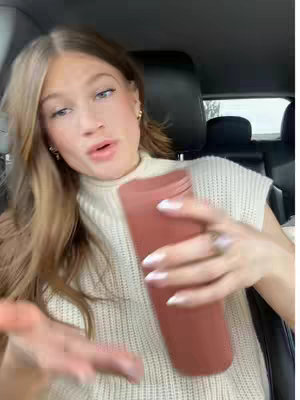 A young woman with long blonde hair is seated in a car, looking towards the camera and speaking. She holds a large, muted rose-colored tumbler or water bottle. She is wearing a cream-colored sweater vest over a high-necked white top, and has earrings. The car's interior, including the seat and headrest, is visible.