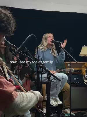 The image captures a live music performance with a female singer as the focal point. The singer, with light blonde hair, is seated on a stool, singing into a microphone. She wears a blue hoodie and jeans. Behind her, a band is visible, and stage equipment like microphones and amps are present. Soft lighting and a dark background create an intimate atmosphere.