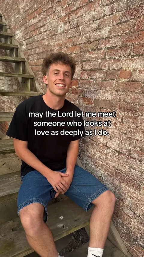 A young man in a black t-shirt and jean shorts sits on wooden stairs. The background is a brick wall. Text overlayed on the image.
