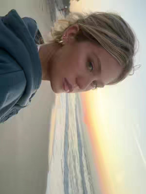 A side-profile shot of a woman with blonde hair, looking toward the left. She’s wearing a blue hoodie with her face near the top left. She has subtle makeup, and the background shows an ocean meeting a colorful sky.