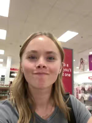 A selfie-style shot taken inside a store. The main element is a young woman with blonde hair looking towards the camera with a slightly amused expression. The background includes shelves with merchandise and signs.