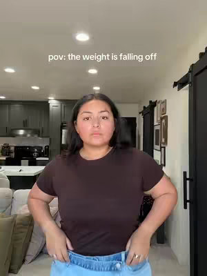 A woman stands in a domestic setting, likely a living room. She is wearing a brown t-shirt and light-wash jeans. The text overlay reads "pov: the weight is falling off." The woman's expression appears neutral or slightly concerned.