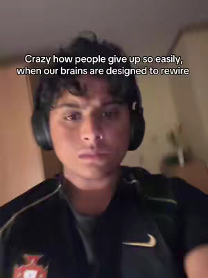 The cover image is a selfie-style shot of a young man indoors. He's wearing a black jacket with a Nike logo and possibly a sports emblem on the left, and black headphones are around his neck. The background appears to be an interior setting, somewhat blurred. Overlaid text at the top reads, 'Crazy how people give up so easily, when our brains are designed to rewire'.