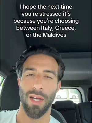 A medium shot features a man sitting inside a car. He is looking directly at the camera and speaking, against a backdrop of what appears to be a sunny day. He has dark hair and a beard, and is wearing a white t-shirt.  Text overlayed above the man reads, "I hope the next time you're stressed it's because you're choosing between Italy, Greece, or the Maldives".