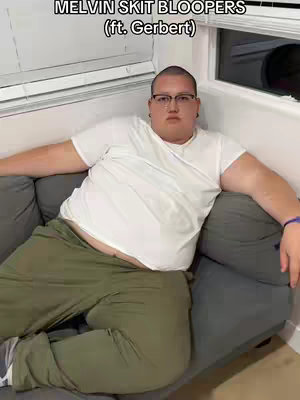 A man with glasses sits on a gray couch, his arms resting along the back. He's wearing a white t-shirt, olive green pants, and appears to be in a living room setting with a window.