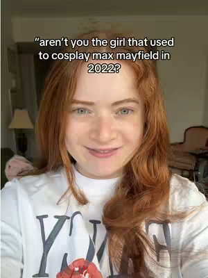 A young woman with long red hair smiles directly at the camera. She is wearing a white graphic t-shirt. The background is slightly blurred, suggesting a home interior with visible elements such as a table lamp, and a corner of a room. The upper portion of the image has overlayed text inside of a rectangle.