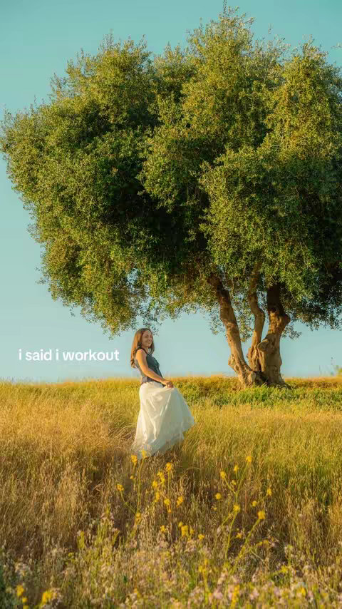 A woman stands seemingly in a field with tall grass and yellow flowers, under a large tree, with a blue sky above. She is wearing a dark top and white skirt, and the image has a warm, natural feel.