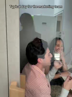 The cover image shows a clear view of three individuals in an office setup. A man in a checkered shirt is looking to his left, partially turned towards the camera, while a woman is seen next to him, laughing, and partially obscured behind the glass window. The background appears to be a well-lit office space with neutral-colored walls. A person from the background is shadowed through the window, which seems to be the central element of the layout in a way.