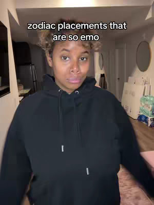 A woman wearing a black hoodie stands in a room, looking at the camera with a slightly serious expression. The room has a neutral color palette with a hint of pink. Text overlay states:'zodiac placements that are so emo'