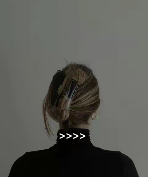 A woman's back is shown, viewed from behind, in a close-up shot. She wears a black turtleneck sweater. Her hair is styled in a bun held by a dark barrette, and gold hoop earrings are visible. The background is a plain, neutral gray.