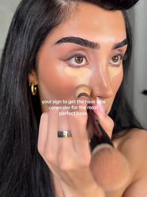 A close-up shot of a woman applying makeup. She is holding a makeup brush to her nose and has concealer applied below her eyes. Text overlayed on the image prompts viewers to get the Haus Labs concealer. The image is well-lit with a focus on the woman's face and the makeup application.