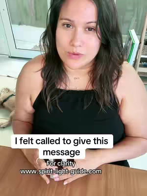 A woman with long dark hair is centered in the frame, looking directly at the camera and speaking. A white rectangular box with black text is overlaid onto the bottom of the image. Behind her, out of focus, is a room with a dog laying on the floor. The background visible behind her is outside.