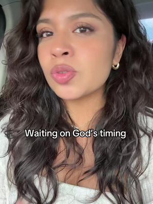 A selfie-style shot featuring a woman with dark, curly hair, looking towards the camera. She has a slight smile and is wearing a white top and gold hoop earrings. The background appears to be inside a car, with the focus primarily on her face and upper body. Overlayed text at the bottom of the image reads: 'Waiting on God's timing'.