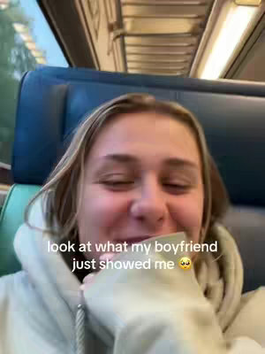 A young woman is seated on a train, facing the camera and partially covering her mouth with her hand. The background shows the interior of the train with seats and overhead compartments. Text overlayed on the image reads: look at what my boyfriend just showed me, followed by a crying face emoji.