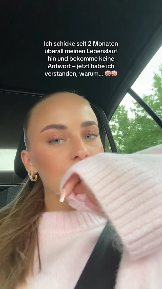 A young woman with pale skin and blue eyes is in a car with the driver's seat in view. She is wearing a pink sweater and has long, manicured nails. Text overlayed on the image in German details her job search frustration. 