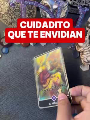 The image shows a hand holding a tarot card with an illustration and text. A red banner with white text is positioned at the top. The background includes various objects and a dark surface.