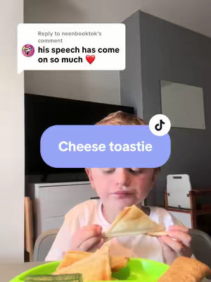 A young boy is seated at a table, holding a triangular cheese toastie in his hands and looking towards the viewer. A green plate with a pickle and more toasties sits in front of him. A speech bubble above the boy says “his speech has come on so much”. A blue rectangular banner with white text that says “cheese toastie” is overlaid on the image.