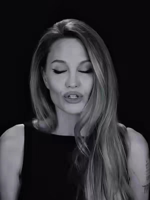 A black and white image featuring a woman with long hair and wearing a black top. She is looking directly at the camera with her mouth open, with closed eyes. The background is plain black.