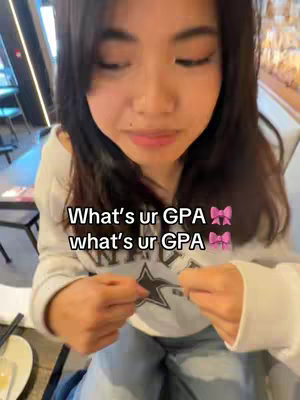 A young woman is positioned in the center, angled from the chest up, looking slightly down toward her hands. She wears a white sweatshirt and is seated at a table. The background is blurred, suggesting a restaurant or cafe setting. Text over the image asks viewers about their GPA.