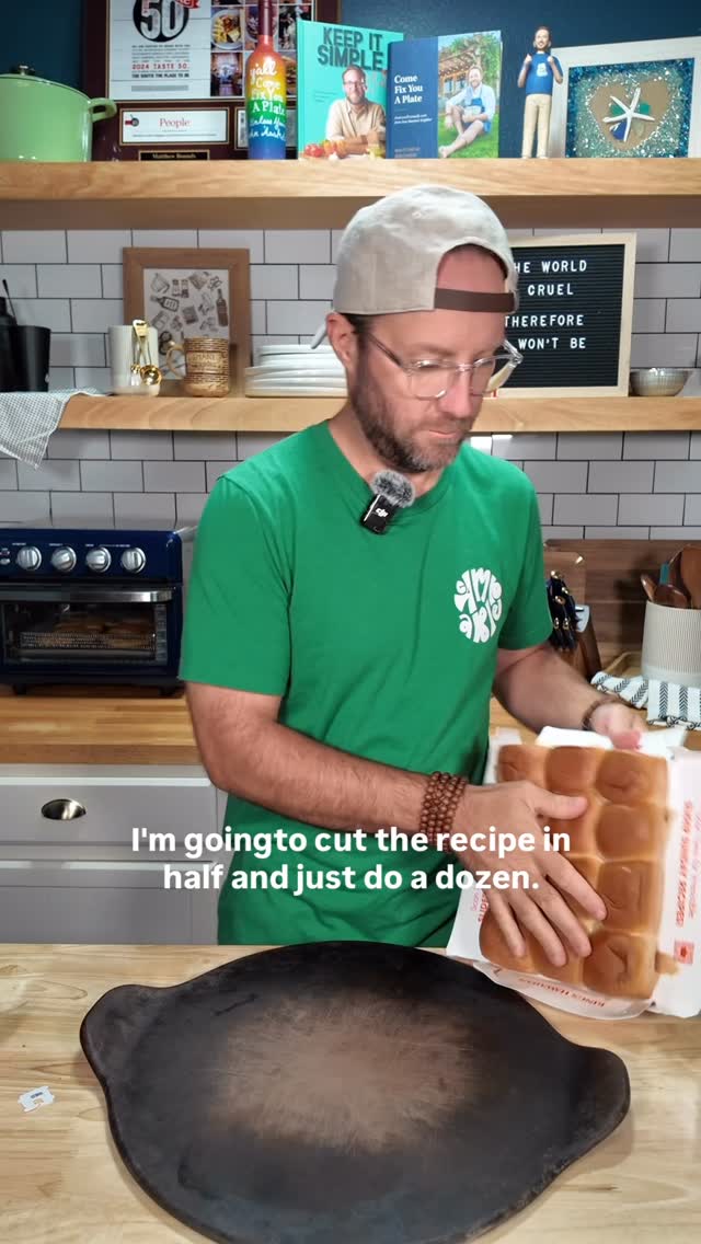 A man in a green shirt and a baseball cap stands behind a wooden counter in a kitchen. He's holding a package of Hawaiian rolls with text overlaid that reads, "I'm going to cut the recipe in half and just do a dozen." A dark, circular object sits in the foreground, likely a pizza stone. Shelves with books, awards, and kitchenware are visible in the background.