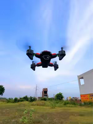 A drone is suspended in mid-air against a backdrop of a clear, light blue sky. The drone, predominantly black with red accents, has visible propellers that suggest motion. Below, there's a green field and a few distant buildings, including one with a prominent white wall and a sign or banner. The overall image is bright and airy.