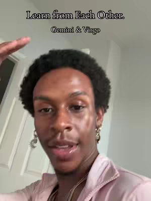 A selfie-style shot featuring a man with curly black hair. The man has an earring, a necklace, and is wearing a light pink shirt. Above the man's head is a text overlay stating, "Learn from Each Other. Gemini & Virgo."