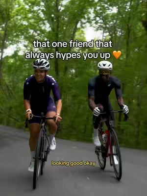 Two cyclists riding bicycles on a paved road, framed by lush green trees on either side. The cyclist on the left is wearing a purple cycling kit, and the one on the right is wearing a black outfit. Text overlays appear on the image.