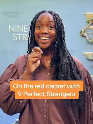 A woman with dark hair and locs smiles at the camera, holding her right index finger up. She wears a buttoned shirt, and an orange rectangle sits in the lower portion of the image with white text. The background is a light blue wall with the words 'NINE STR' written in white.