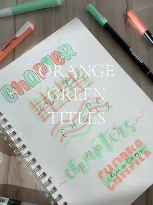 A notebook page with various handwritten title ideas for chapters, displayed in orange and green markers against a blurred background of matching pens, markers, and a wooden surface.