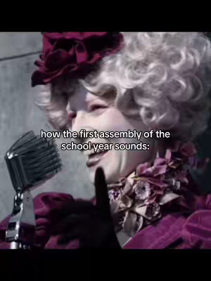 The image features a person, dressed in a theatrical costume and makeup, holding a microphone and speaking. Text overlayed on the image reads 'how the first assembly of the school year sounds:'. The background and attire suggest a theme related to 'The Hunger Games.'