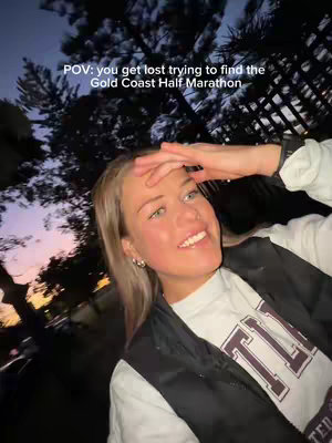 A young woman is smiling, looking towards the right of the frame with her hand on her forehead. The background is out of focus, with greenery and a blurred, colorful sunset forming a backdrop. The woman is wearing a white t-shirt with a logo and a black vest.