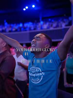 A man with his arms raised high in praise during a worship session. The background shows a large crowd of people in an auditorium, lit with blue and purple lighting. Text overlay is featured on the image.