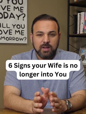 The cover image features a man in a light blue button-down shirt looking directly at the camera. Overlayed on him is a white rectangular text box with the title '6 Signs Your Wife Is No Longer Into You' in black text. The background appears to be a neutral-toned wall, with a sign with writing on it and a bookshelf visible to the left and right of the man.