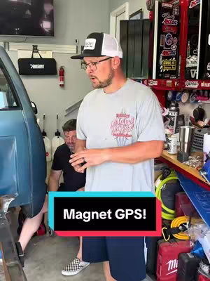 The cover image shows a man in a garage setting. The man, wearing glasses, a backward baseball cap, and a light gray t-shirt, is the central focus, gesturing with his hands and speaking. Behind him, another man is partially visible, working on what appears to be a car. The background includes garage tools, shelves, and equipment. The foreground features part of the car, and text in a colored box states 'Magnet GPS!'