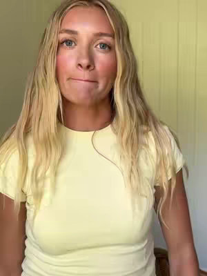 A woman with long blonde hair and blue eyes looks directly at the camera with a slightly pursed mouth. She is wearing a light yellow shirt, and the background is a light, neutral color. The lighting is soft and natural.