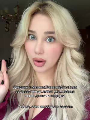 A selfie-style image featuring a fair-skinned woman with long, wavy blonde hair. She has a surprised expression with wide, light blue eyes and slightly parted lips. Her cheeks are flushed with pink blush. She is pointing her right index finger upwards, and her left hand is out of view. The background is blurred, but hints at an interior space, possibly a room. Text overlays the image in Russian.