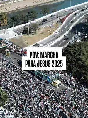 An aerial view of a large crowd of people gathered on a street, with a river and bridge with traffic in the background. A white rectangular overlay with black text is positioned in the lower center, and a yellow truck is driving down the street. The overall scene is bathed in sunlight.