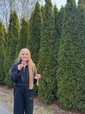 A woman with long blonde hair stands in front of a row of tall, green, cone-shaped trees. She wears a black sweatshirt and black pants and holds a small object in her hand. Her expression is pleasant.