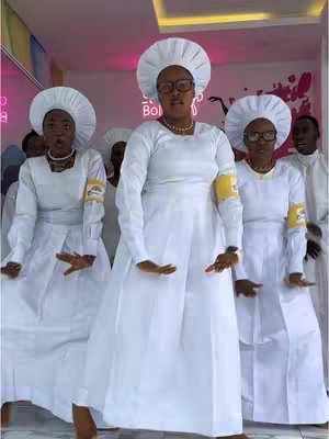 The image shows a group of individuals dressed in white, performing what appears to be a dance. The group is comprised of several people, all wearing white dresses, hats, and glasses. They appear to be on a stage or in a well-lit room, with the background of pink and magenta hues .
