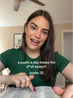 A woman with brown hair and a green shirt is smiling with a book in front of her. Text at the bottom center reads 'a psalm a day keeps the d*vil away!!! psalm 25.'
