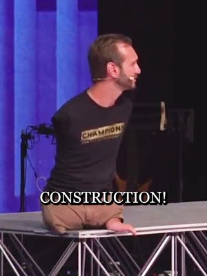 A man with no arms stands on a stage, facing to the right. He is wearing a black t-shirt with gold lettering that says CHAMPION. The bottom part of his legs are visible, he is wearing tan pants. The background is blurred with purple and blue lights. The text 'CONSTRUCTION!' is overlaid at the bottom of the image.