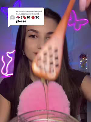 A woman is centered in the frame, with a wooden honey dipper held close to the camera. Honey is dripping from the dipper onto a pink fluffy item. The background suggests a softly lit room with artificial lighting in the shape of butterflies. A small text bubble with some text and emojis is visible in the upper left corner.