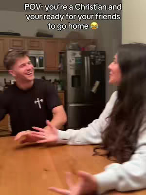 Two people sitting at a wooden table, inside a kitchen. The foreground features a man in a black t-shirt with a cross design. The text reads "POV: you're a Christian and your ready for your friends to go home".