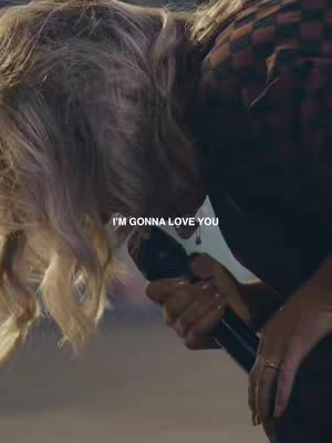 A person singing into a microphone, with blonde hair partially obscuring their face. The background is softly lit, and the person is wearing a patterned plaid shirt and has rings on their hand. The overlaying text 'I'm gonna love you' reinforces the emotional expression.