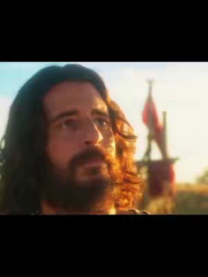 The image shows the upper portion of a man, presumably Jesus, with medium-length dark hair and a beard. He has a contemplative expression, looking slightly upwards and to the right. The lighting suggests a sunny or outdoor setting, with the light catching his hair and beard, giving them a sense of glow. In the background, a mast with red banners is visible on the right.