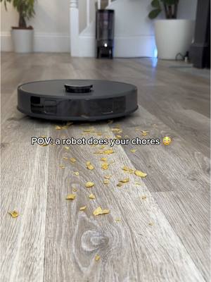 A robot vacuum cleaning up scattered corn flakes on a wooden floor with a staircase and white potted plants in the background.