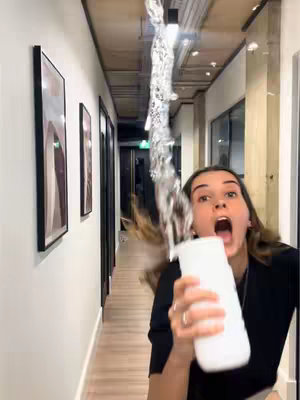 A woman in a black top reacts with an open mouth as a stream of water shoots toward her, originating from an unseen point above. She holds a white bottle. The shot is inside a hallway with side and back-lit lighting through a series of doors.